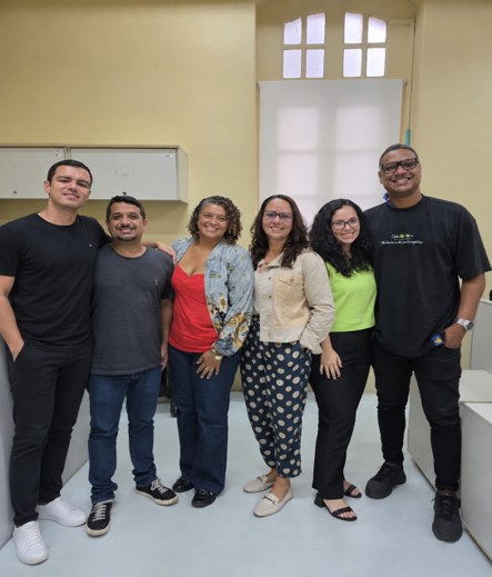 Fotografia de grupo mostra seis pessoas sorrindo, reunidas em sala de aula ou ambiente corporativo, possivelmente para registrar equipe ou encontro. Ao fundo, aparecem quadro branco, tela de projeção, janelas altas e paredes claras; participantes usam roupas casuais em cores variadas.