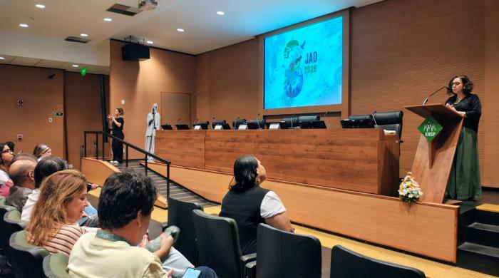 imagem retrata o auditório da ENSP na abertura da JAD 2026, com um telão trazendo a logo do evento, uma mulher apresentando e pessoas na plateia.